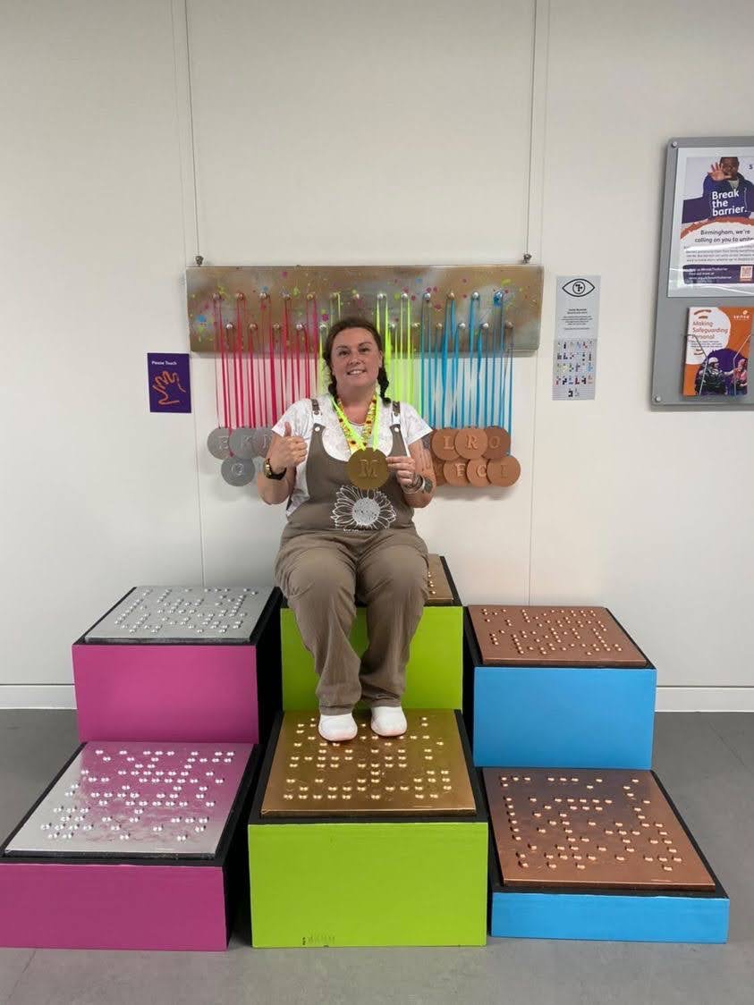 A person sitting on the top podium out of 6 podiums with braille on them holding a gold medal with one of her thumbs up. The medal reads the letter M in non-braille.