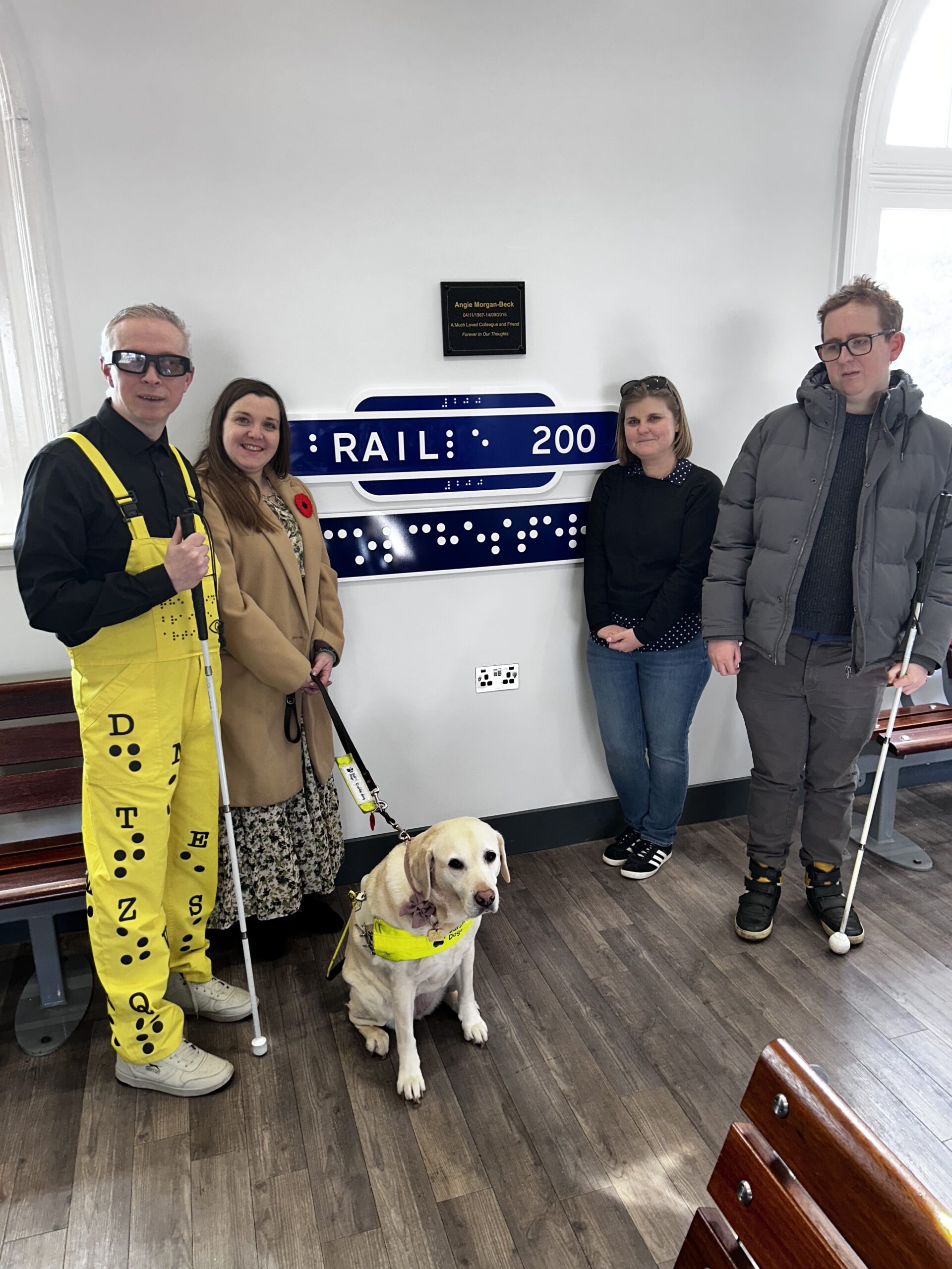 Mr Dot with supporters standing in front of a braille railway sign, one supporter with a guide dog. Mr Dot is wearing a yellow braille outfit.